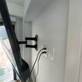 A black TV mounted on an articulating wall bracket in a bright room with white walls, with a black power cable running down to a white electrical outlet below.