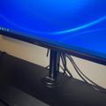 A computer monitor displaying a blue desktop background, mounted on a black stand with cables visible, positioned above a laptop keyboard on a desk