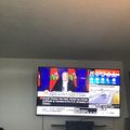 Television mounted on wall showing news broadcast with person speaking at podium with Canadian flags in background