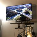 Large flat screen TV mounted on wall bracket displaying aerial view of highway interchange with rivers and autumn foliage, with visible cables and mounting hardware below