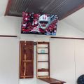 Interior room with mounted TV showing football game, wooden shelving unit, bench, and corrugated metal ceiling