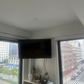 A flat-screen TV mounted on a white wall above a wooden dresser in a modern apartment, with large windows showing an urban cityscape view on either side.