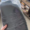 Person holding a dark foam mattress or cushion wrapped in clear plastic packaging in a home office setting