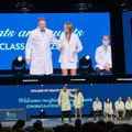 Medical school graduation ceremony with students in white coats on stage receiving recognition, with large screen displaying ceremony details above
