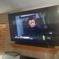 A flat-screen TV mounted on a wall showing a scene from what appears to be a TV show or movie, with a person in dark clothing visible on screen. The TV is positioned above a white tiled fireplace, and there are wooden blinds visible on the left side of the room.