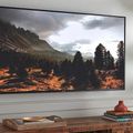 Wall-mounted TV displaying mountain landscape scene above wooden console table with books and decorative items, with plant visible on right side