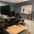 A modern home office workspace with a standing desk setup, computer monitor, ergonomic chair, and colorful artwork on gray paneled walls