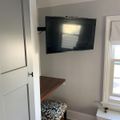 A small room with a wall-mounted TV above a wooden desk shelf, featuring a patterned chair, white door, and window with radiator