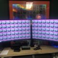 A dual monitor computer setup on a wooden desk displaying repeated anime-style character images as wallpaper, with a collection of framed pictures or cards visible on the wall above