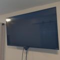 A large flat-screen TV mounted on a beige wall with white curtains visible to the left and cables hanging below the TV