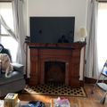 Living room with wooden fireplace mantel, flat screen TV mounted above, gray armchair, hardwood floors, white curtained windows, and various items scattered on the floor