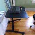 A black height-adjustable desk with wheels holds an open laptop computer in a room with green walls and hardwood flooring