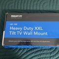 A Mount-It brand Heavy Duty XXL Tilt TV Wall Mount box for 60-120 inch TVs, sitting on a green quilted surface with colorful floral fabric visible at the top