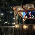 Outdoor evening setup with wooden pergola structure, string lights, and mounted TV screen showing a person, surrounded by trees and fencing