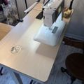 A sewing machine setup on a light wooden desk with a task lamp, thread spools, and sewing supplies in a craft room workspace
