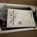 A black device or equipment piece in white foam packaging inside a cardboard box, with instruction manual and hardware bag visible on top