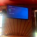A TV screen displaying a user profile selection menu with options for Eric Messenger, Jess, Rune, and New Profile, mounted on a red wall in an indoor setting with windows and plants visible.