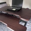 A laptop computer sits on a wooden desk with an adjustable mechanism, positioned on a bed with gray bedding in a bedroom setting.