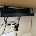 Under-desk cable management system with black Mount-It! cable tray mounted beneath wooden desk surface, with various cables and power adapters connected to wall outlets