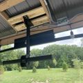 A black mechanical lift or hoist system mounted to wooden ceiling beams in what appears to be a garage or workshop, with a view of a green lawn and trees visible through the open doorway