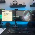 A dual monitor gaming setup with RGB lighting, mechanical keyboard, and floating shelves displaying collectibles and headphones