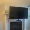 Living room with wall-mounted TV above white fireplace, decorative candles on shelf, and large plant in corner