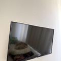 A flat-screen television mounted on a white wall, displaying a blurred image. Below the TV are two yellow and red signs or posters partially visible at the bottom of the frame.