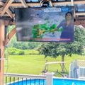 Outdoor TV mounted under wooden gazebo structure showing weather forecast, with swimming pool and yard visible in background