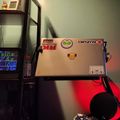 A desk setup with a laptop covered in stickers, red LED lighting underneath, a monitor on the right, and shelving with small figurines on the left