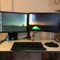 A dual monitor computer setup on a white desk with a keyboard, mouse, and drink can, displaying desktop wallpapers of mountain landscapes