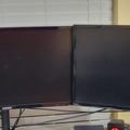 Two black Samsung computer monitors on a glass desk with white tiled wall in background