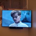 A large flat-screen TV mounted on a wall displays a scene with a young person in a white jacket, with decorative stairs visible to the left side of the frame