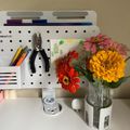 A white pegboard desk organizer with office supplies and colorful zinnia flowers in a glass vase on a white desk surface