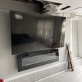 A large flat-screen TV mounted on a wall above a dark floating shelf in an unfinished basement or construction area with exposed ceiling tiles and concrete floors