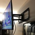 A large flat-screen TV mounted on an adjustable black wall bracket displaying a blue-tinted image, with additional TV mounting hardware visible on the white wall