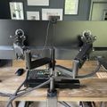 A dual monitor desk setup with two computer screens mounted on adjustable arms above a wooden desk, with various office supplies and equipment visible in a home office environment