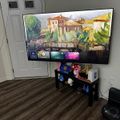 A wall-mounted TV displaying a colorful painted landscape scene with a menu interface, positioned above a black entertainment stand with decorative items in a corner room with striped carpeting.