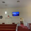 Interior of a church or chapel with white walls, red carpeted pews, pendant lighting, a wall-mounted TV screen, and an exit sign visible