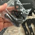 A person's hand holding a small plastic bag containing black metal mounting hardware and brackets. Additional black metal mounting pieces and a black mounting plate with "MOUNTIT!" text are visible on a textured surface in the background.