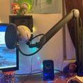 A desk setup with a computer monitor, microphone on an adjustable arm, small speaker, and decorative figurines on a shelf in the background