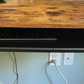 A wooden desk with a black keyboard tray underneath, showing cables and wall outlets below the work surface