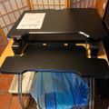A black adjustable standing desk converter with two tiers - an upper desktop surface and lower keyboard tray - positioned in front of wooden room divider screens