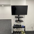 A home gym setup with a wall-mounted TV above floating shelves and a dumbbell weight rack below containing various weights and exercise equipment