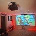 A game cave room with red neon lighting, displaying a football game on a large projection screen, with gaming equipment and furniture visible.