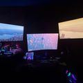 A dark room with multiple large screens displaying various colorful images and videos, with computer equipment and gaming setup visible below the monitors