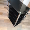 A modern black glass TV stand with multiple shelves and chrome metal support poles, positioned on a wooden floor in a home interior