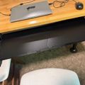 A home office setup with a wooden desk shelf above a black adjustable keyboard tray, featuring a laptop, mouse, and cables on a carpeted floor