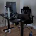 A modern home office setup with a black standing desk, ergonomic office chair, computer monitor, laptop, and various cables on a wooden floor against gray walls
