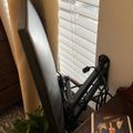 A surfboard leaning against a wall next to window blinds, with a bicycle and a book titled "Samuel Adams" visible in the foreground