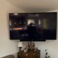 A large flat-screen TV mounted on a white wall above a wooden dresser with decorative items, showing what appears to be a video game or media content on the screen.